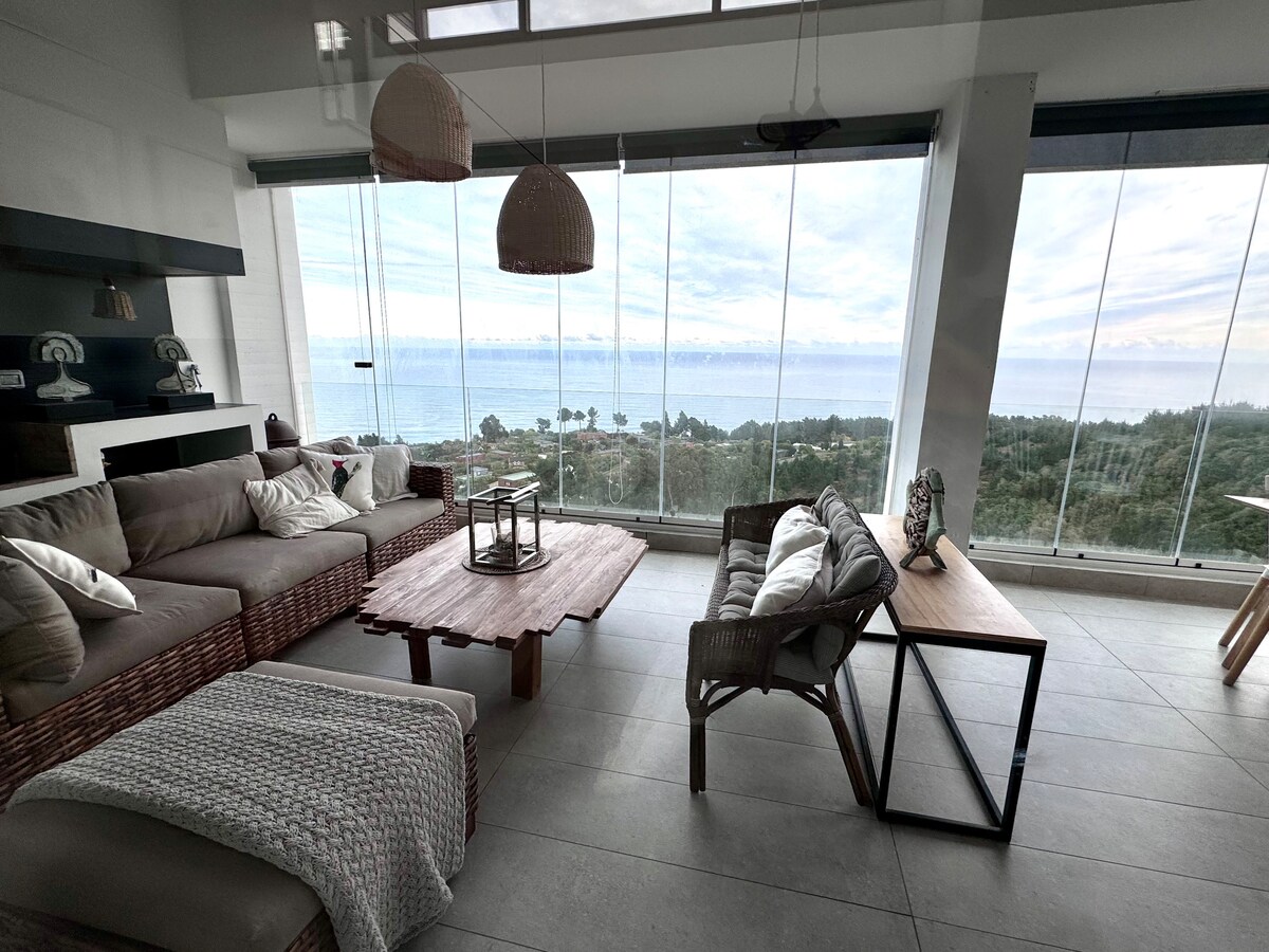A modern living area features large glass windows offering panoramic views of the coastline. Natural light floods the space, highlighting a neutral color palette with a comfortable sofa, a wooden coffee table, and decorative elements that enhance the open and airy atmosphere.