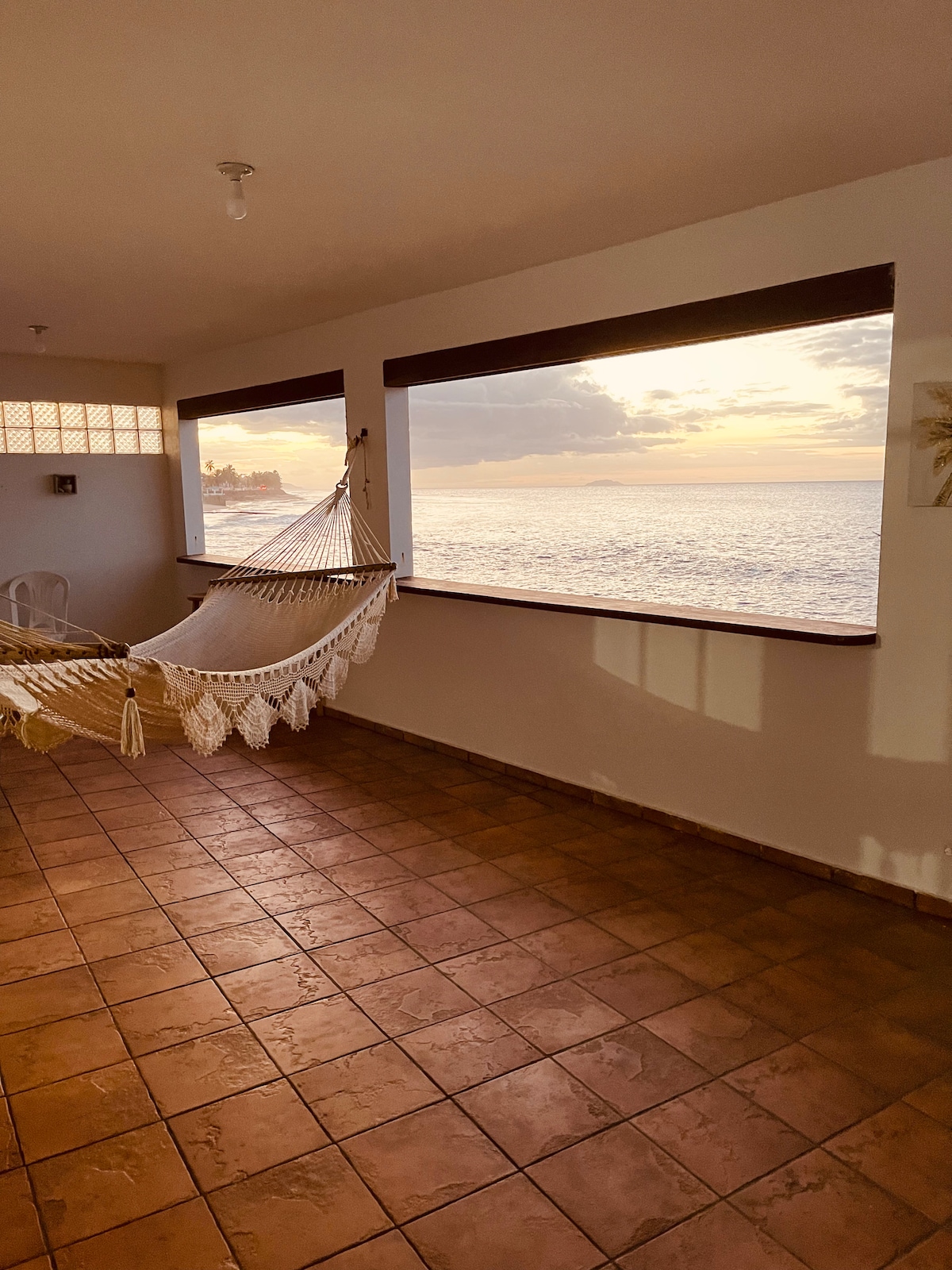A cozy seating area is highlighted by a large window offering ocean views. A hammock is suspended nearby, inviting relaxation. The tiled floor and light walls create a spacious ambiance, while soft natural light filters in, enhancing the serene environment.
