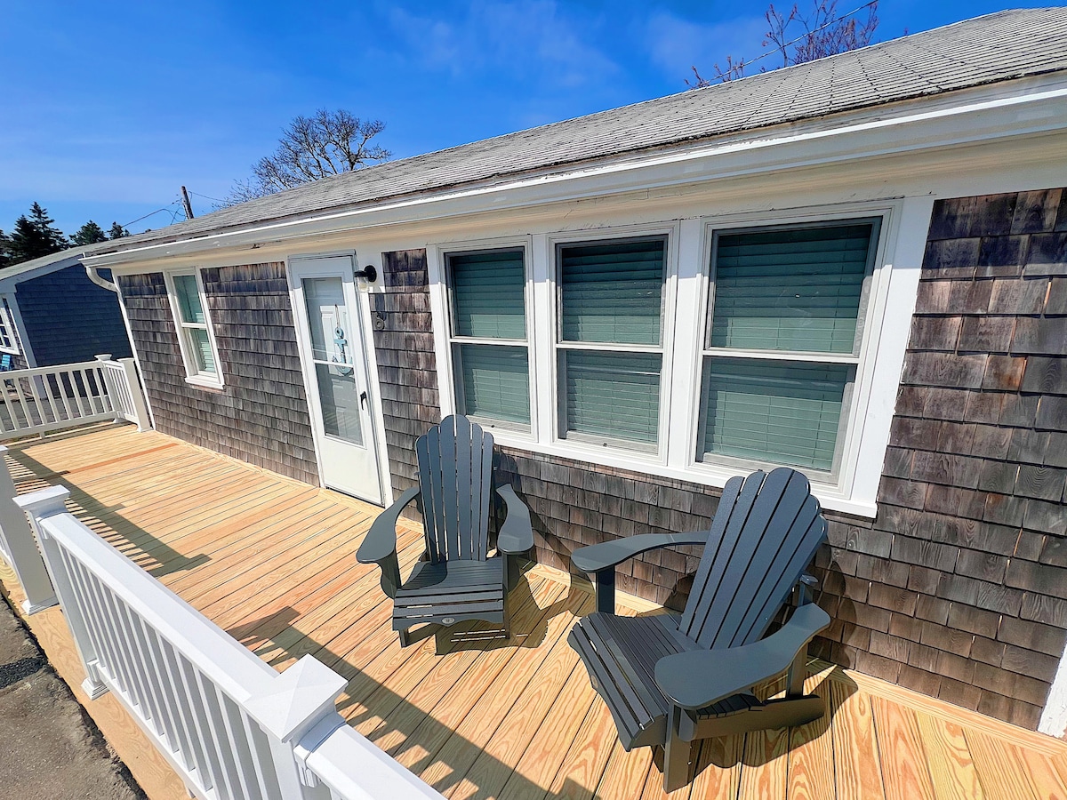 A wooden deck features two classic Adirondack chairs, positioned to enjoy the surrounding views. The cottage's exterior showcases shingled siding and four windows allowing natural light. A door leads inside, framed by fresh, inviting outdoor space.