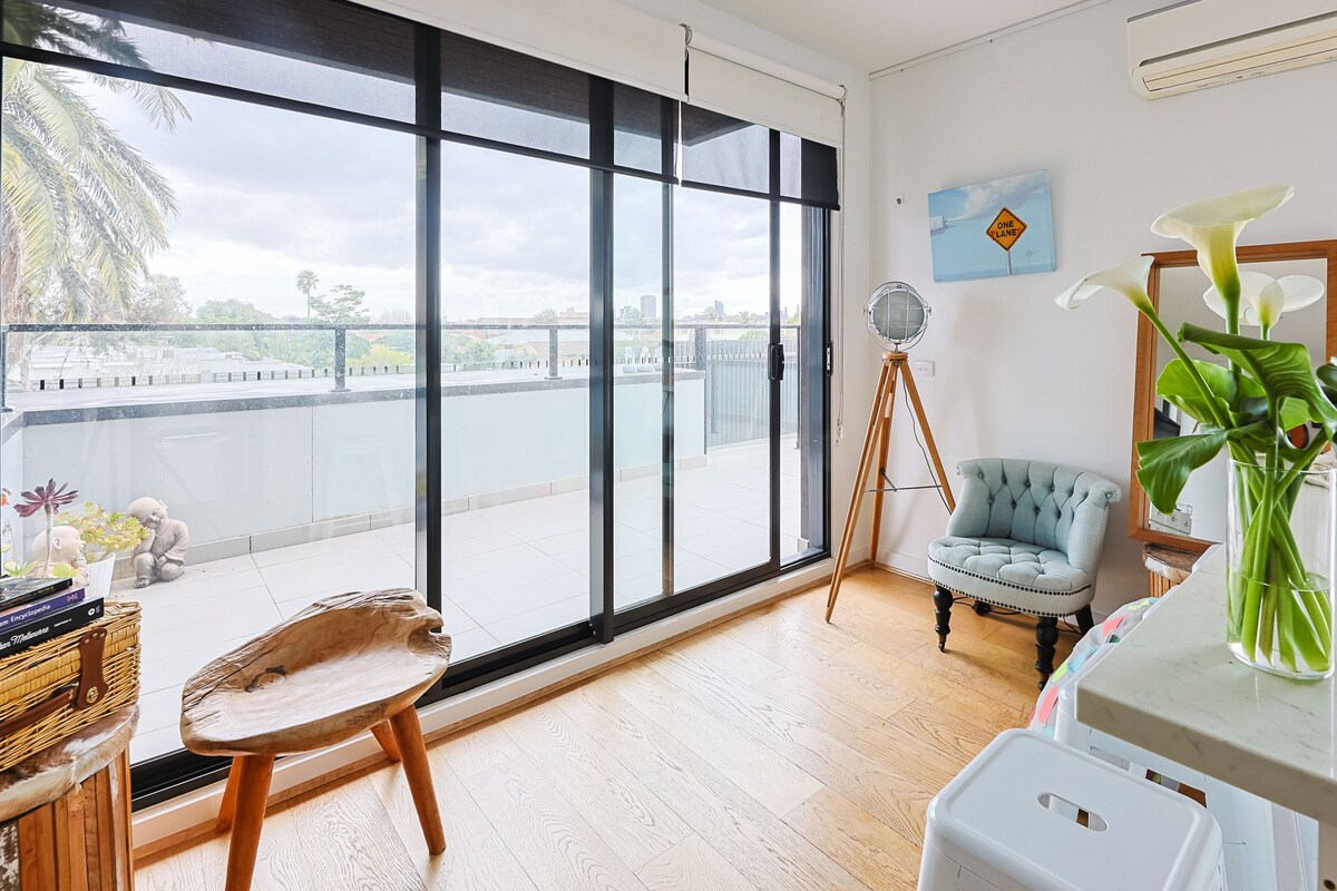 A sunlit living area features large sliding glass doors that open to a spacious private balcony. A stylish, light blue armchair is positioned near the windows, complemented by a unique wooden side table. Light wooden flooring enhances the room's modern aesthetic.