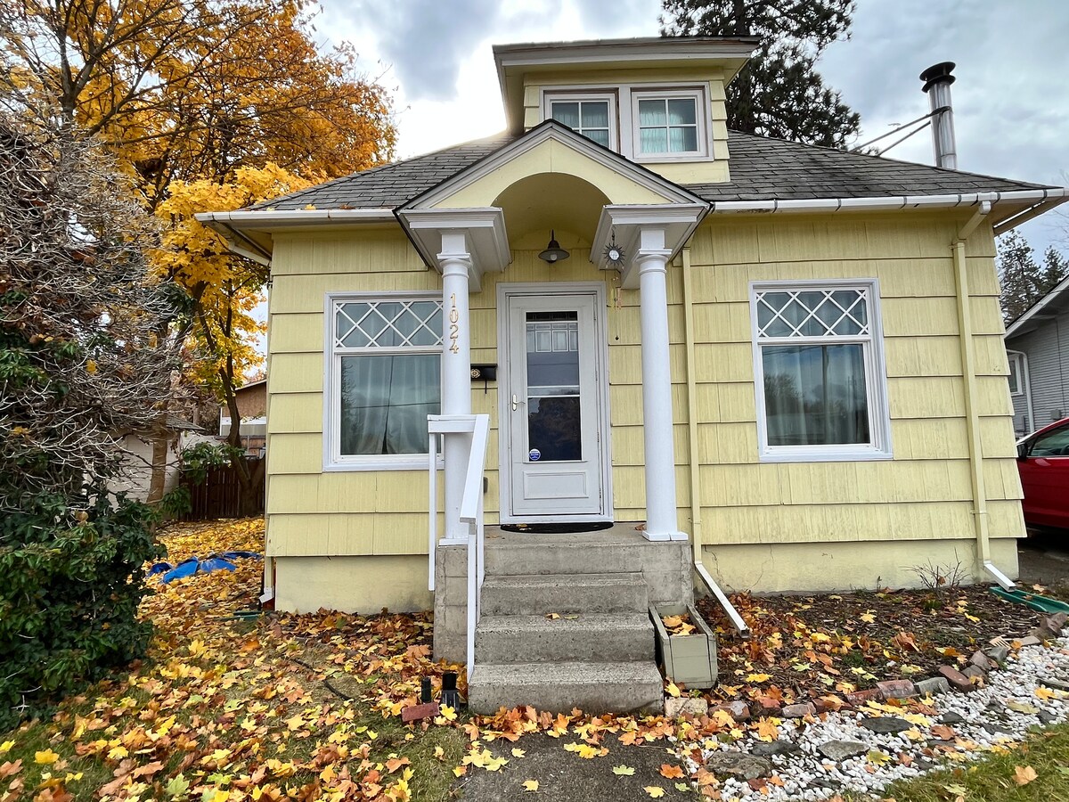 The exterior of a charming yellow house is displayed, featuring a welcoming front porch with white columns. Two large windows with decorative grids contribute to the home's character. Autumn leaves blanket the front yard, and a hint of greenery from nearby trees is visible.