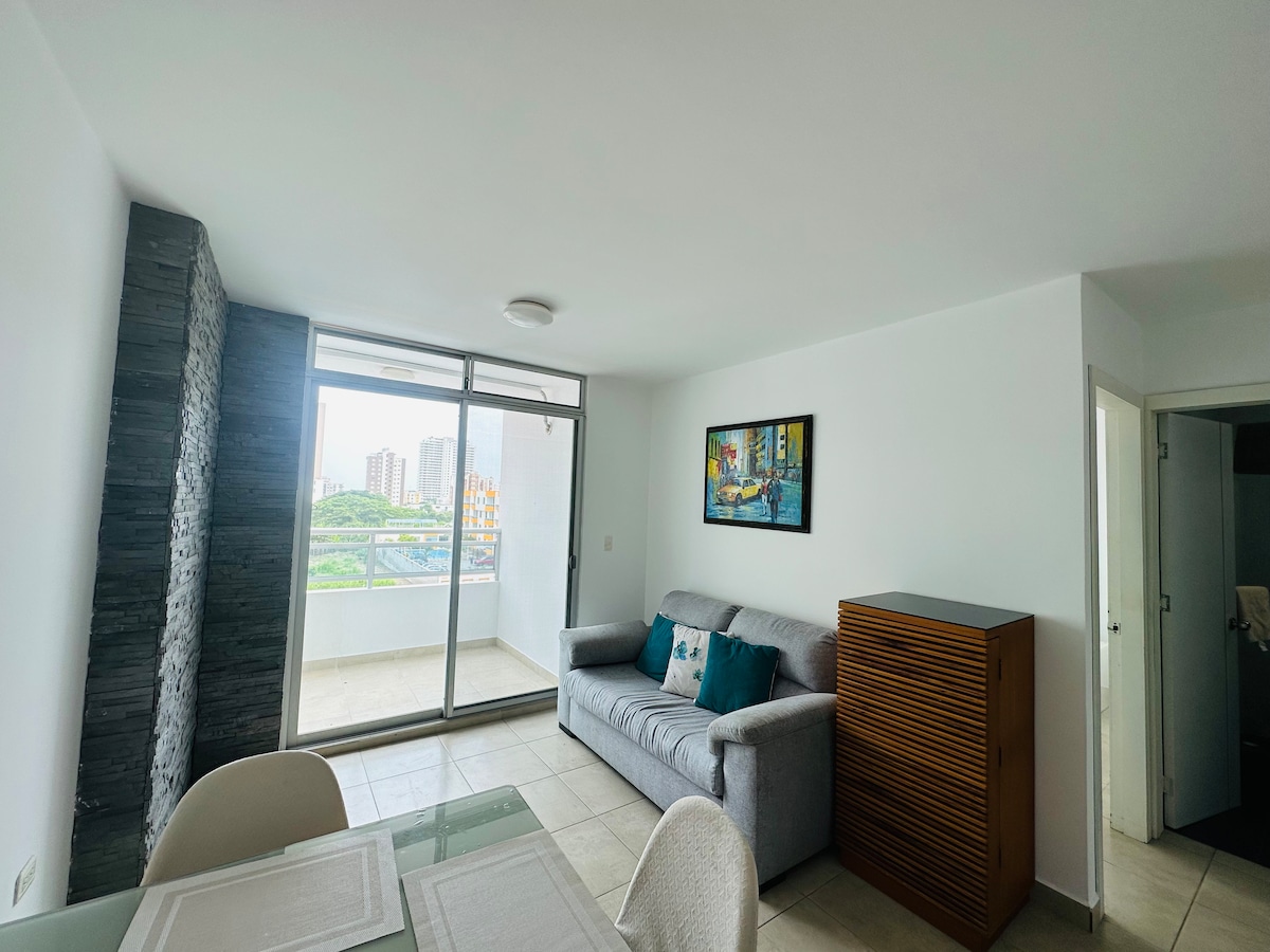 A modern living area is featured with a gray sofa positioned near a large sliding glass door that allows natural light. A colorful wall art piece complements the space, while a wooden cabinet offers additional storage. A dining table is visible to the left.
