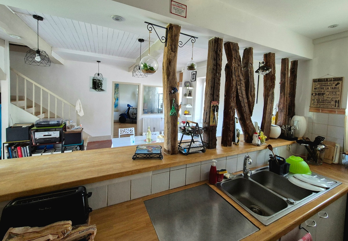 A bright kitchen area features a wide wooden counter with a sink and various kitchen utensils. Natural wood elements enhance the space, while light fixtures hang above. In the background, shelves display decor items, and a glimpse of the living area is visible.