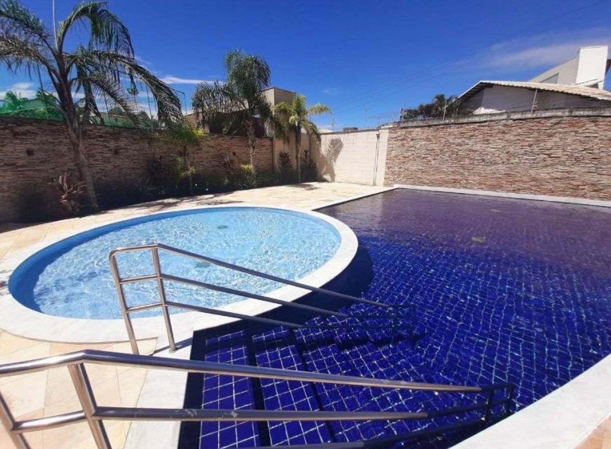 A spacious outdoor pool area is displayed, featuring a large rectangular pool and a circular hot tub. Surrounding plants add greenery, and sunlit tile surfaces enhance the inviting atmosphere of the space.