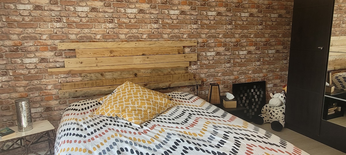 A cozy sleeping area features a wooden headboard against a brick wall. The bed is draped with a patterned blanket, complemented by a decorative yellow pillow. A bedside table is positioned nearby, with a small collection of items visible. A plush toy and a storage basket can also be seen.