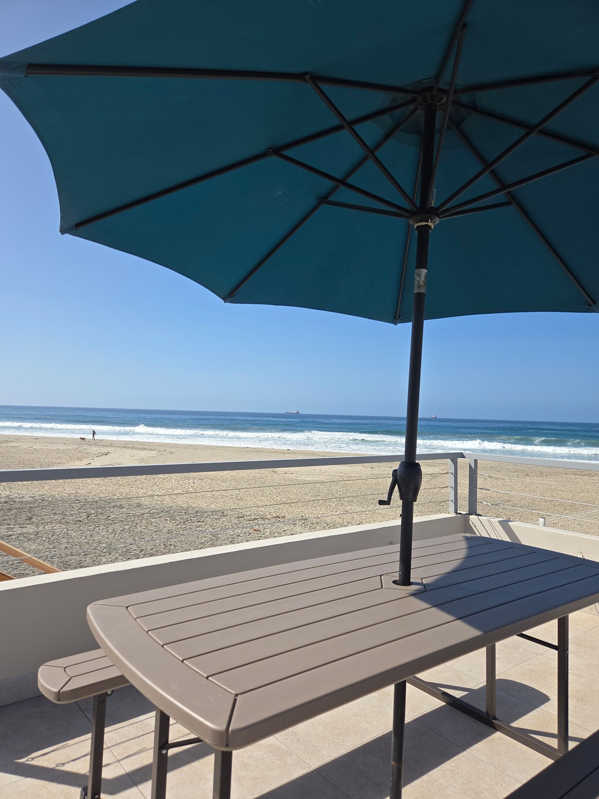 A shaded outdoor table with a teal umbrella is positioned on a patio, offering a clear view of the sandy beach and the ocean waves. The scene captures a sunny day with people enjoying the beach in the distance.