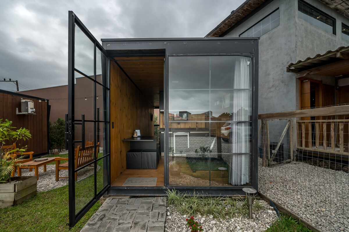 A modern micro-house with large glass doors allows natural light to enter. The exterior features a grassy area and simple landscaping, while a kitchenette is visible inside. The design emphasizes a seamless transition between indoor and outdoor living.