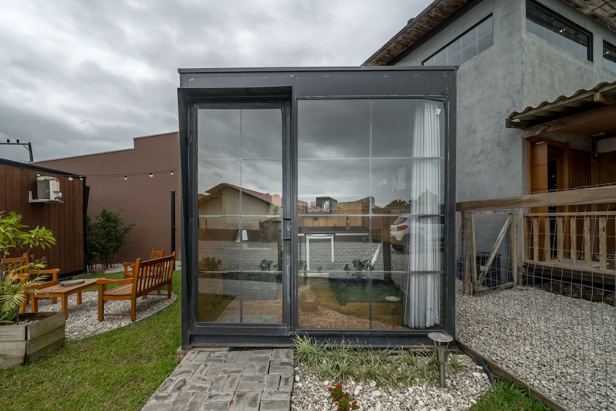 A modern micro house is featured with large glass windows, allowing ample natural light. The building's sleek design contrasts with the surrounding green grass and gravel. A cozy outdoor seating area is visible beside the structure, enhancing the overall appeal of the space.