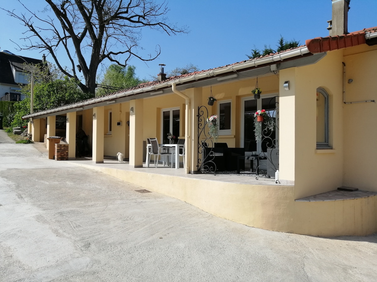 A charming exterior view of the house is presented, highlighting a spacious terrace with outdoor seating. Sunlight illuminates the façade, showcasing neutral-colored walls and a tiled roof. Lush greenery surrounds the property, contributing to a serene setting.