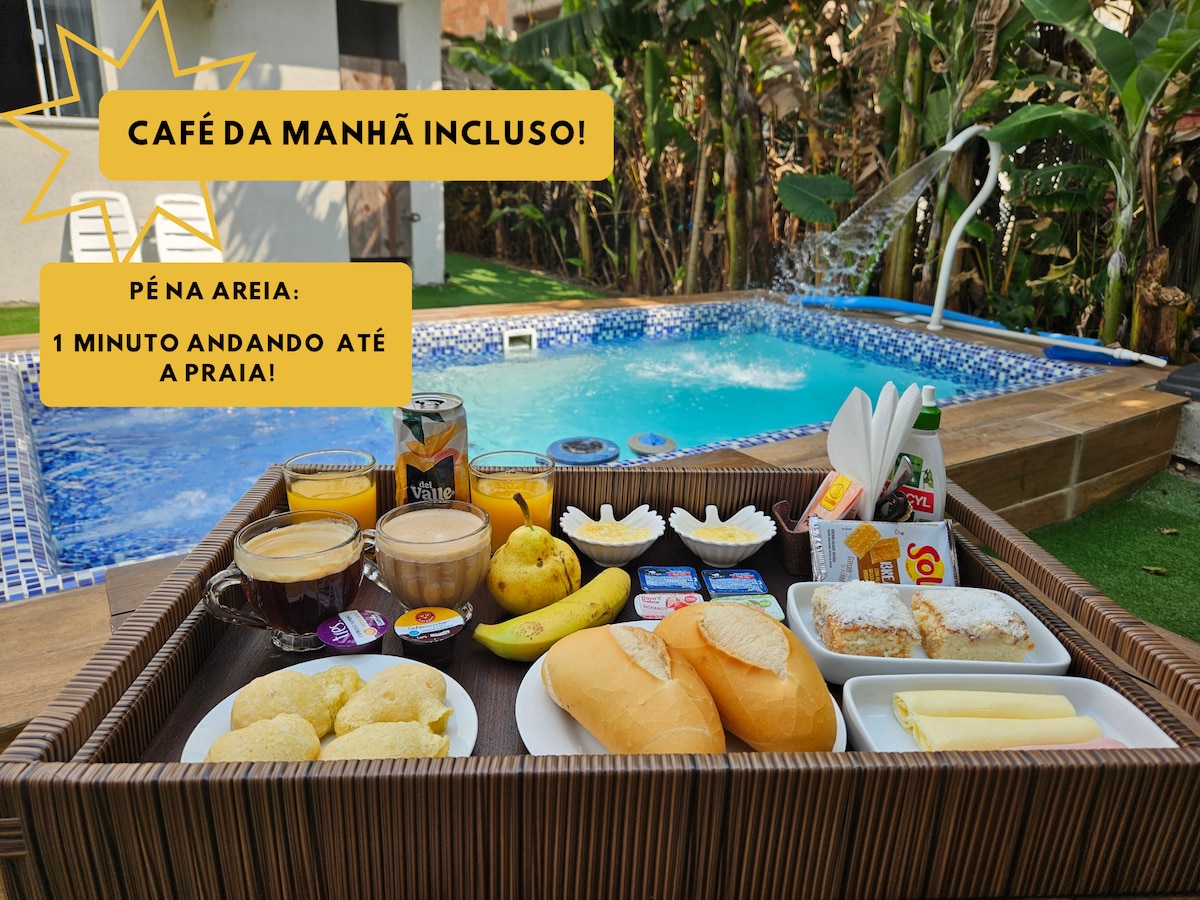 A wooden tray is displayed, filled with a variety of breakfast items including fresh bread, pastries, fruits, and beverages. In the background, a shared pool is visible, surrounded by greenery, highlighting the inviting atmosphere of the outdoor space.
