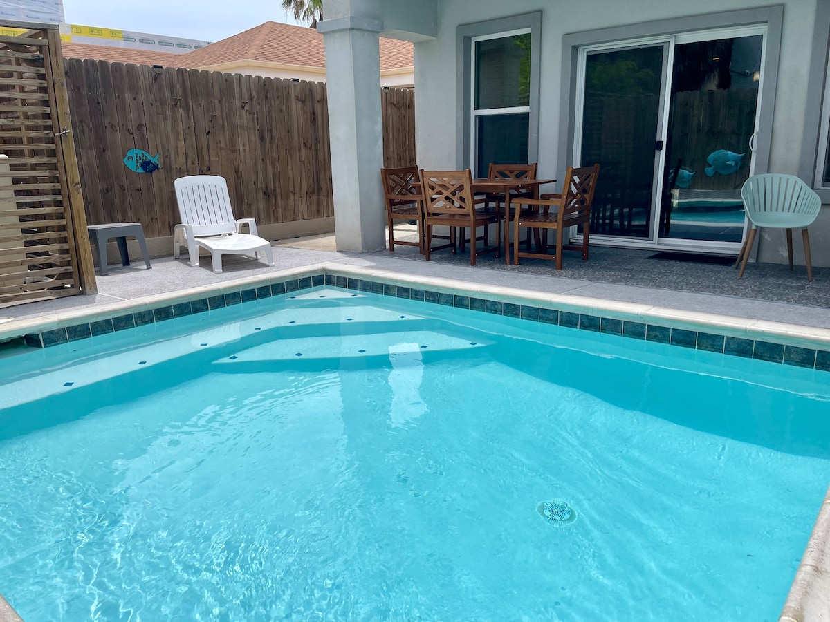 A private backyard features a bright blue heated pool surrounded by a patio area. Nearby, a wooden dining table with six chairs provides an outdoor dining option, alongside a lounge chair for relaxation. A fence creates a sense of enclosure and privacy.