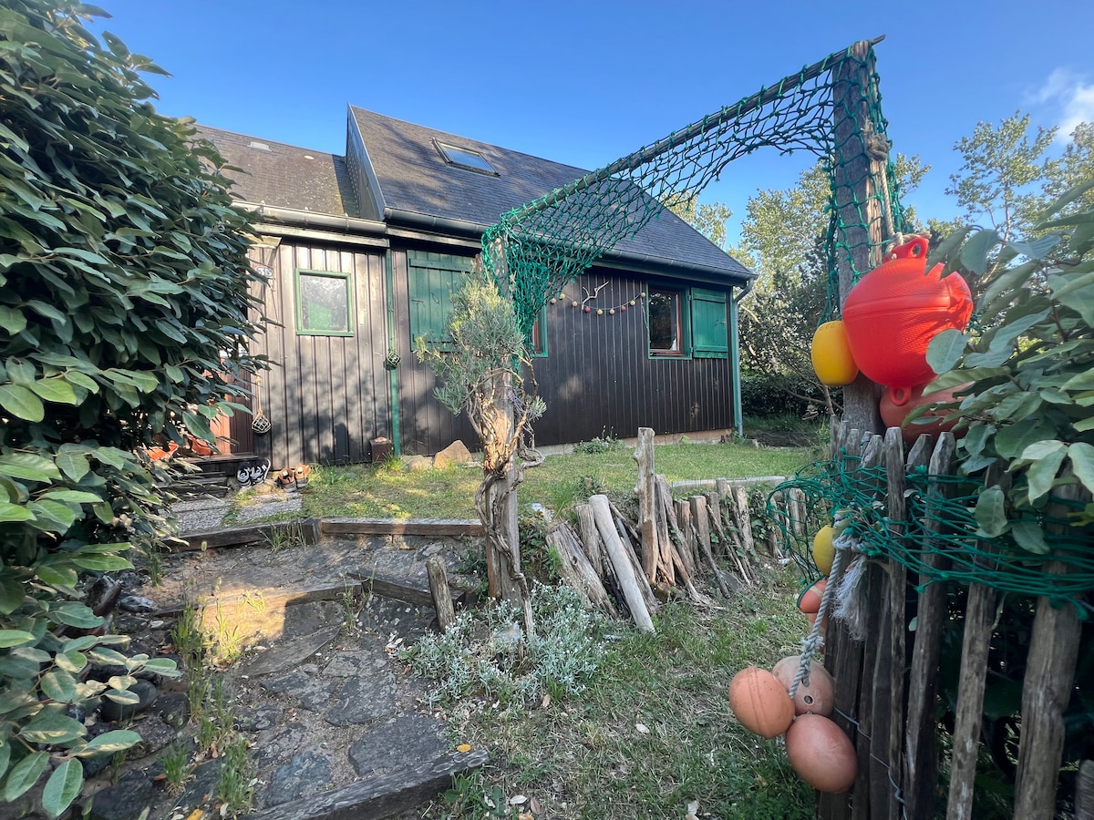 The exterior of a charming chalet is depicted, surrounded by lush greenery. A rustic fence made of timber adds natural texture, while a decorative net hangs with colorful items. Clear skies and soft lighting enhance the welcoming vibe of the entrance.