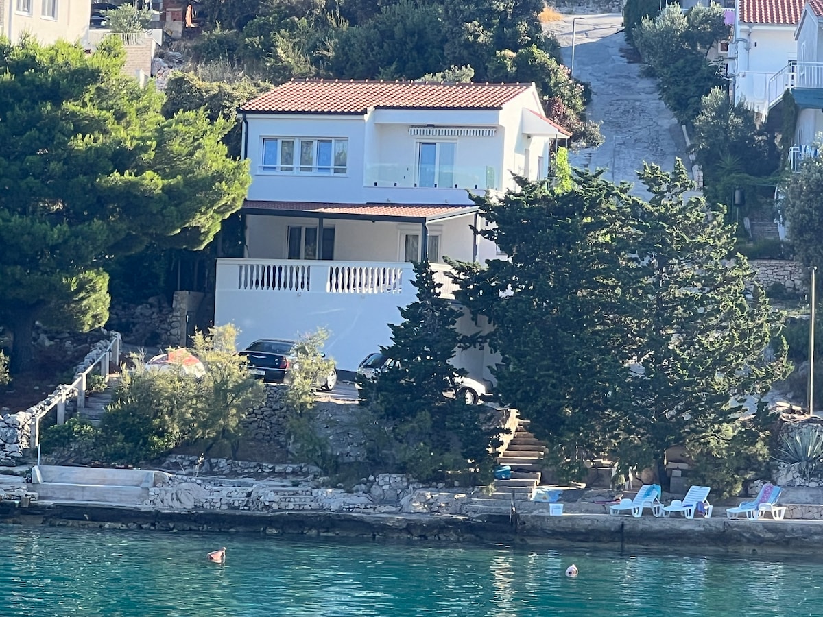 A two-story apartment building is set among lush greenery alongside the tranquil sea. A terrace with railings is visible on the second level, while several parked cars are seen in front. Sun loungers are positioned nearby, offering a space for relaxation by the water.