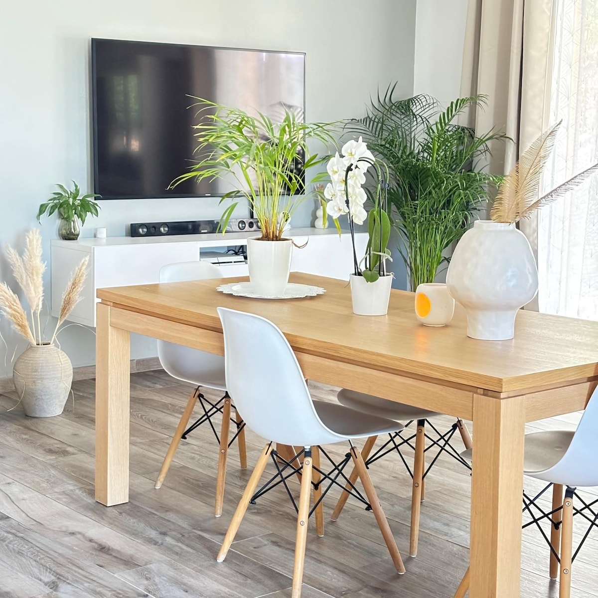 A simple wooden dining table is surrounded by white chairs, featuring light wood legs. Potted plants and a white vase with decorative accents adorn the table. A flat-screen TV is mounted on the wall, complementing the room's bright and airy atmosphere.