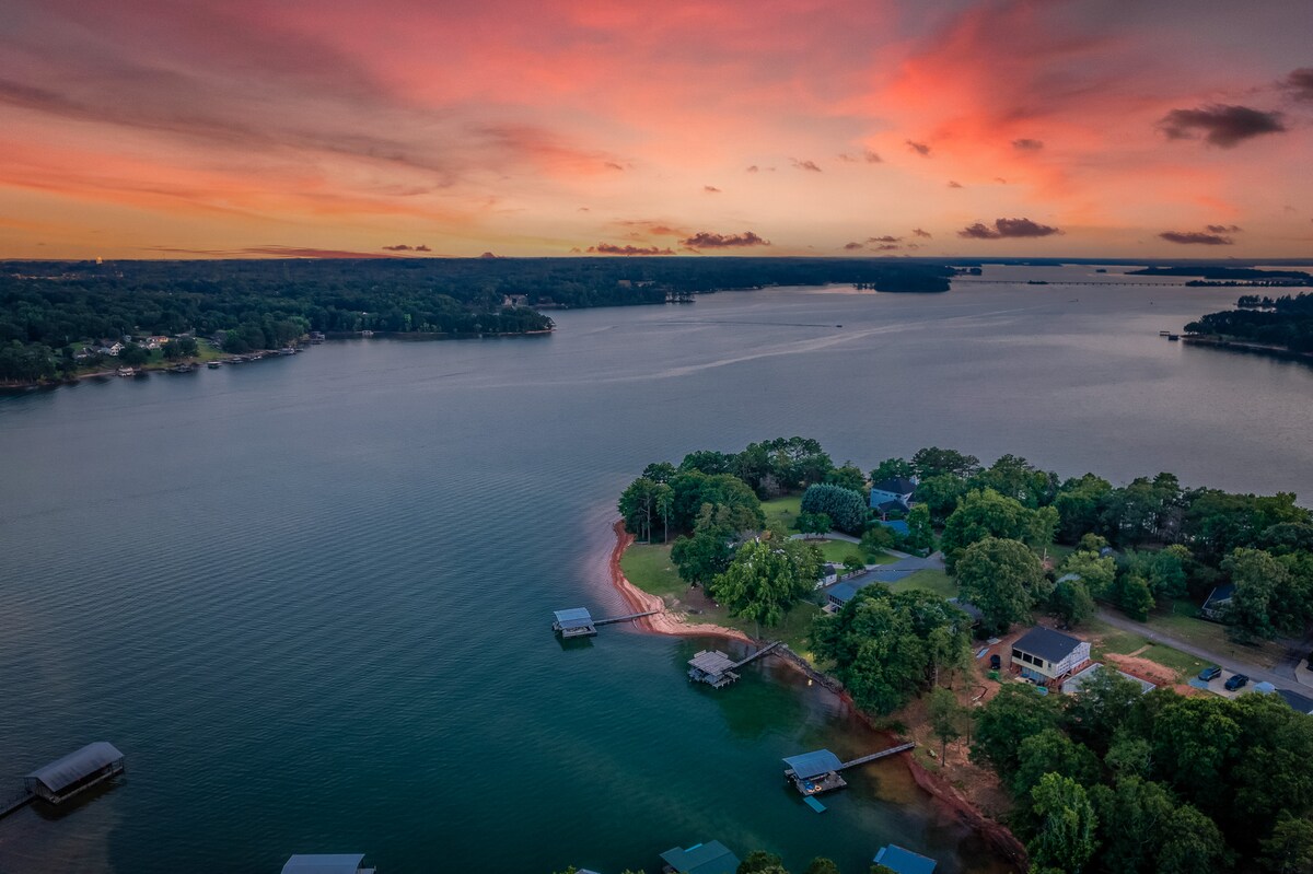 An aerial view captures the calming beauty of Lake Hartwell at sunset, with hues of pink and orange lighting the sky. The shoreline is lined with lush greenery, leading to docks nestled along the water's edge, creating a serene and inviting atmosphere.