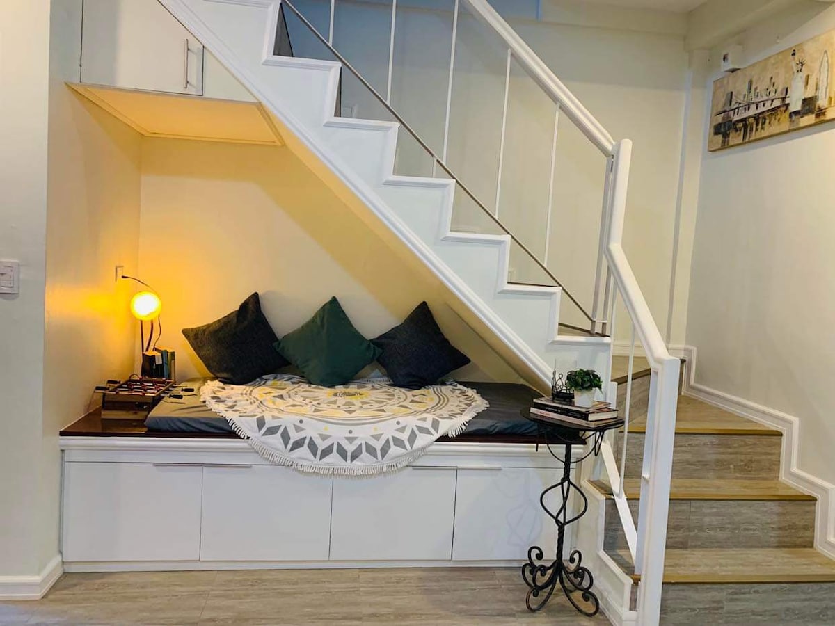 A cozy nook is situated beneath the staircase, featuring a comfortable seating area adorned with plush pillows and a decorative throw. A small side table holds a lamp and decorative items, while the stairway ascends alongside, creating an inviting atmosphere for relaxation.