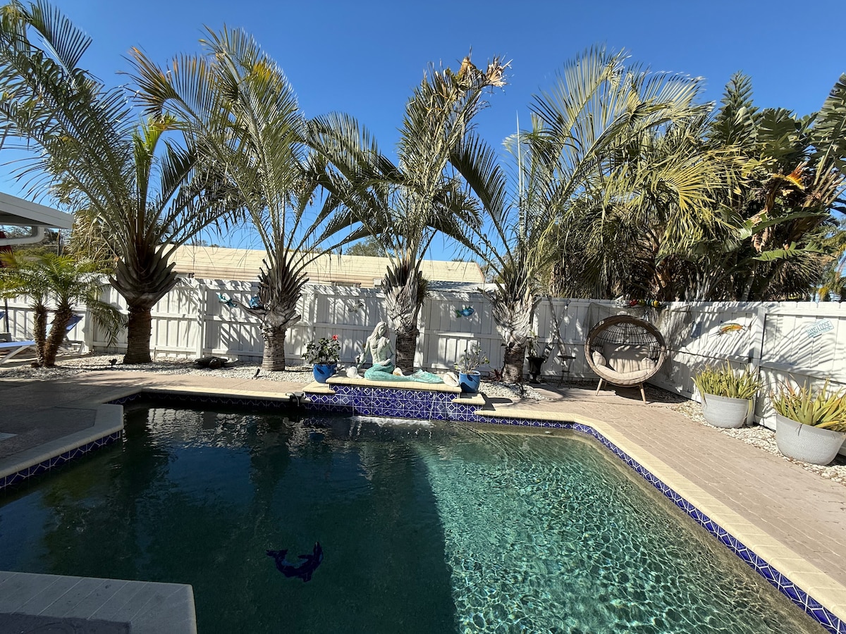 A heated pool is surrounded by lush palm trees, providing a sense of seclusion. A stone statue and decorative elements are featured along the pool's edge. Sunlight reflects off the water, creating a serene atmosphere in this private outdoor oasis.