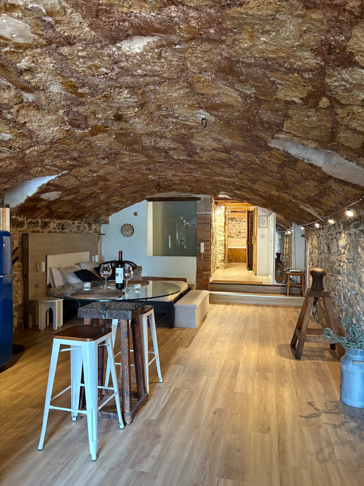 An open living space is presented with a rustic stone ceiling and warm wooden flooring. A dining table is set for two, accompanied by stools. Soft lighting enhances the natural stone walls, leading to an interior hallway that connects to other areas of the home.