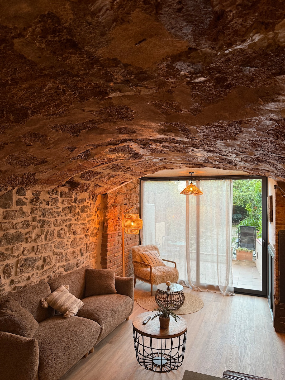 A cozy seating area features a brown sofa and an armchair positioned near a circular table. Natural stone walls and a softly arched ceiling are complemented by warm lighting. Large glass doors open to a lush garden space, enhancing the inviting atmosphere of the room.