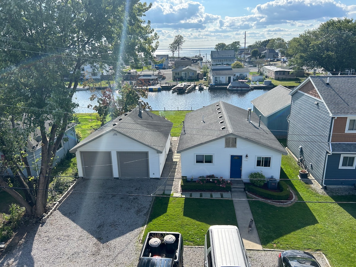 An aerial view showcases a charming cottage adjacent to a peaceful waterway. Two garages are visible, along with a well-maintained yard featuring a path leading to a blue front door. The serene landscape includes several boats in the water and a clear sky dotted with clouds.