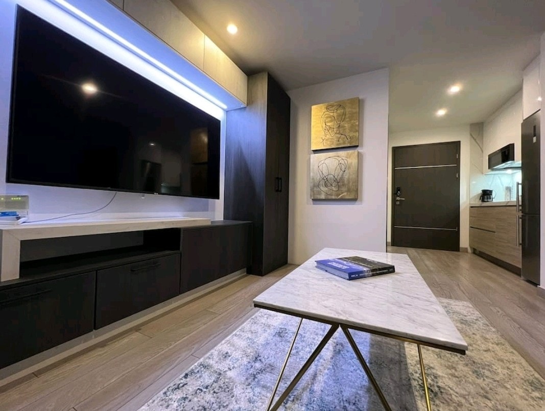 A modern living area is featured with a large flat-screen television mounted on the wall, complemented by minimalist cabinetry. A marble coffee table rests on a textured area rug, while two abstract art pieces are displayed above. The well-equipped kitchen is visible in the background.