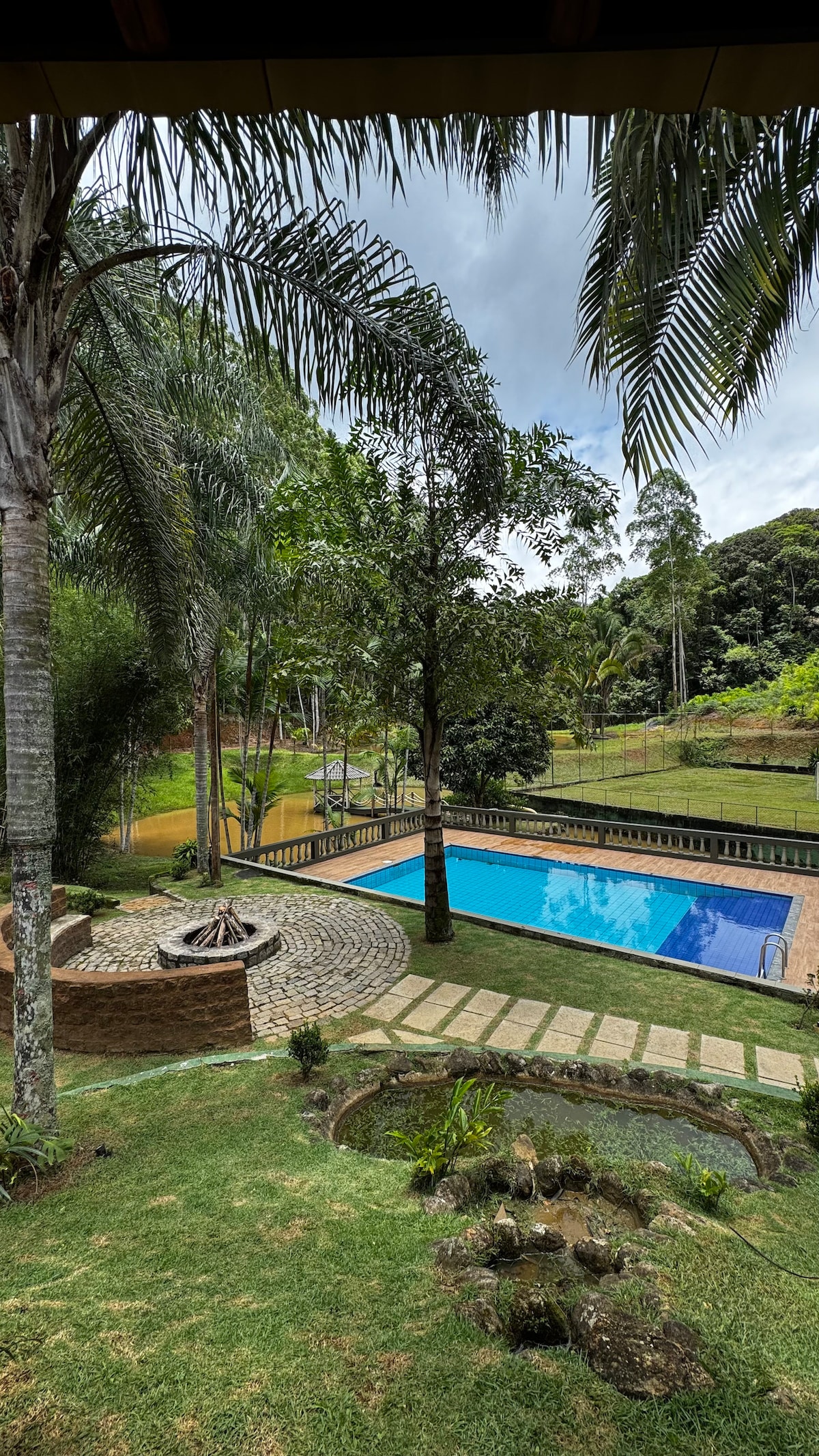 The outdoor area features a spacious swimming pool surrounded by lush greenery. A stone pathway leads to a firepit, creating a designated gathering spot. Tropical trees and hills provide a natural backdrop, enhancing the serene atmosphere of the site.