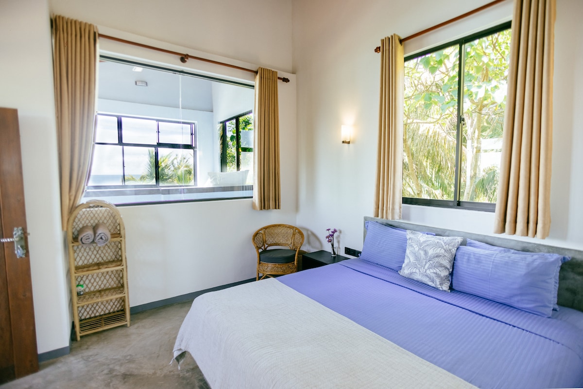 A bright bedroom features large windows that invite natural light and provide views of lush greenery. A spacious bed dressed in soft purple linens is complemented by neutral-toned curtains. A bamboo chair is positioned nearby, enhancing the inviting atmosphere of the room.