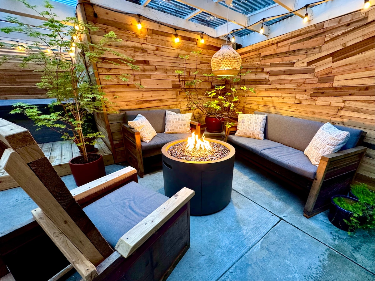 An outdoor living space is highlighted with cozy seating arranged around a circular fire pit. Warm lighting from above creates a soothing ambiance against the wooden wall. Potted plants add a touch of greenery, enhancing the tranquil setting for relaxation.