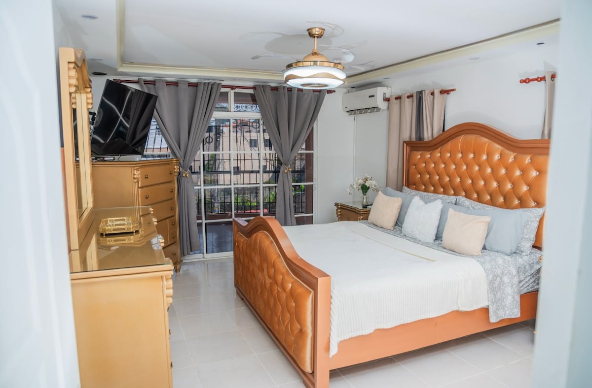 A well-appointed master bedroom features a spacious, tufted bed with light-colored bedding and multiple decorative pillows. A stylish dresser complements the room's elegant design, while sheer curtains frame the large window offering views of the outdoors. A television is mounted opposite the bed.