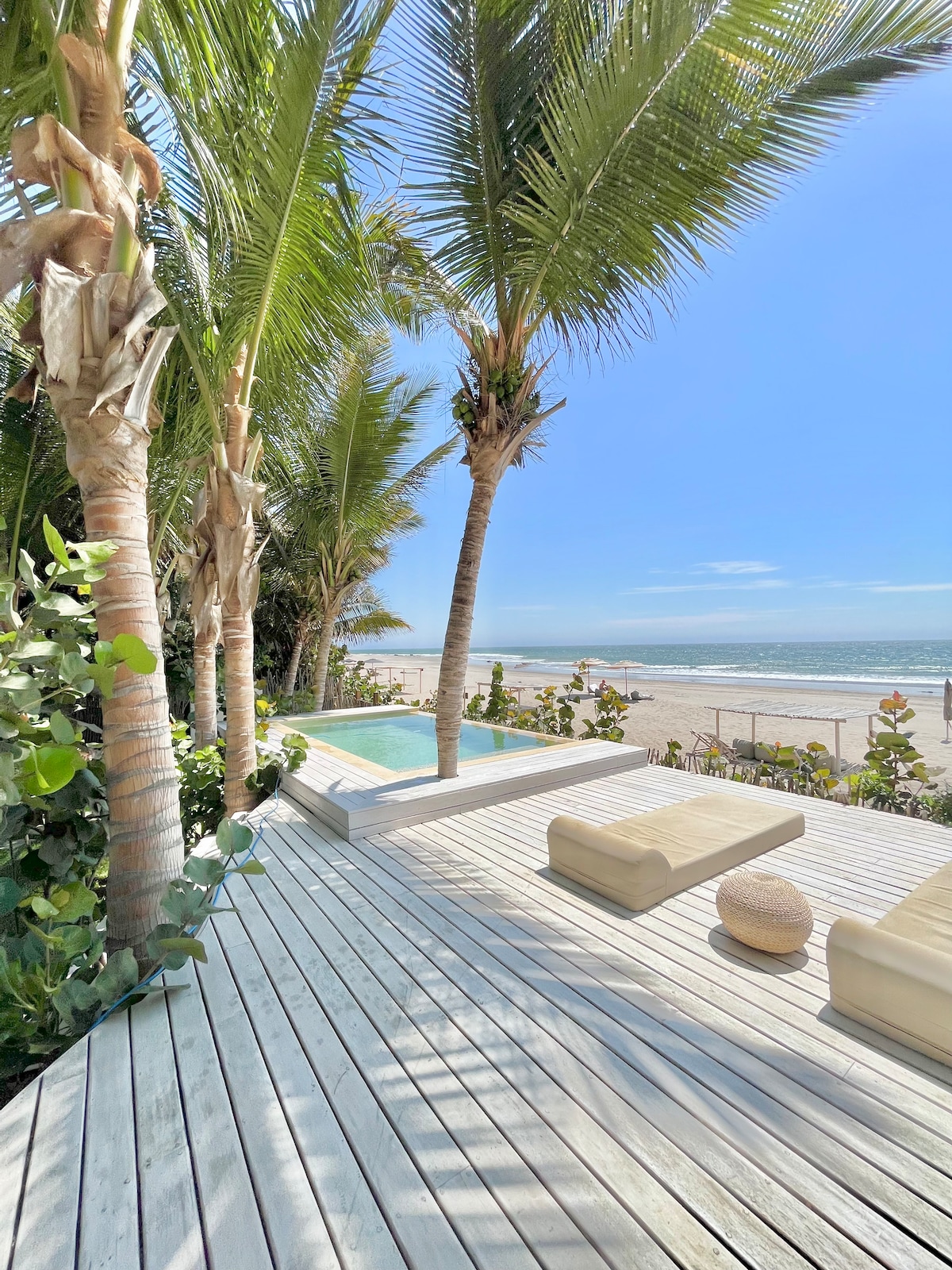 A sunlit deck is surrounded by tall palm trees, leading to a small, clear pool. Two understated lounge chairs are placed nearby, with the expansive beach visible in the background, showcasing golden sand and gentle waves under a clear blue sky.