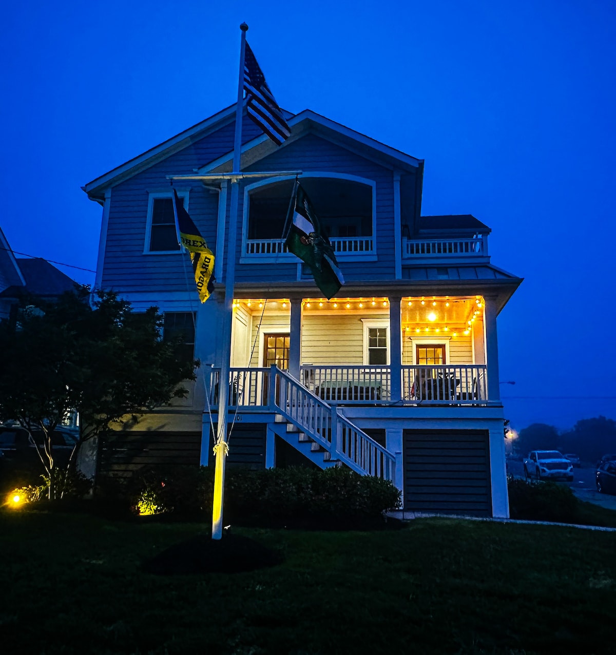 A charming two-story house is depicted at dusk, accentuated by soft outdoor lighting and flags along the porch. The front porch offers ample space for relaxation, while the inviting design showcases a well-maintained lawn and garden, enhancing the welcoming atmosphere.