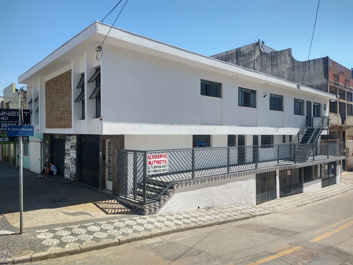 A modern building featuring a white facade with stone accents. The structure includes multiple windows and a balcony area with a railing. A pathway runs along the front, bordered by a patterned stone surface. The surrounding area shows urban characteristics.