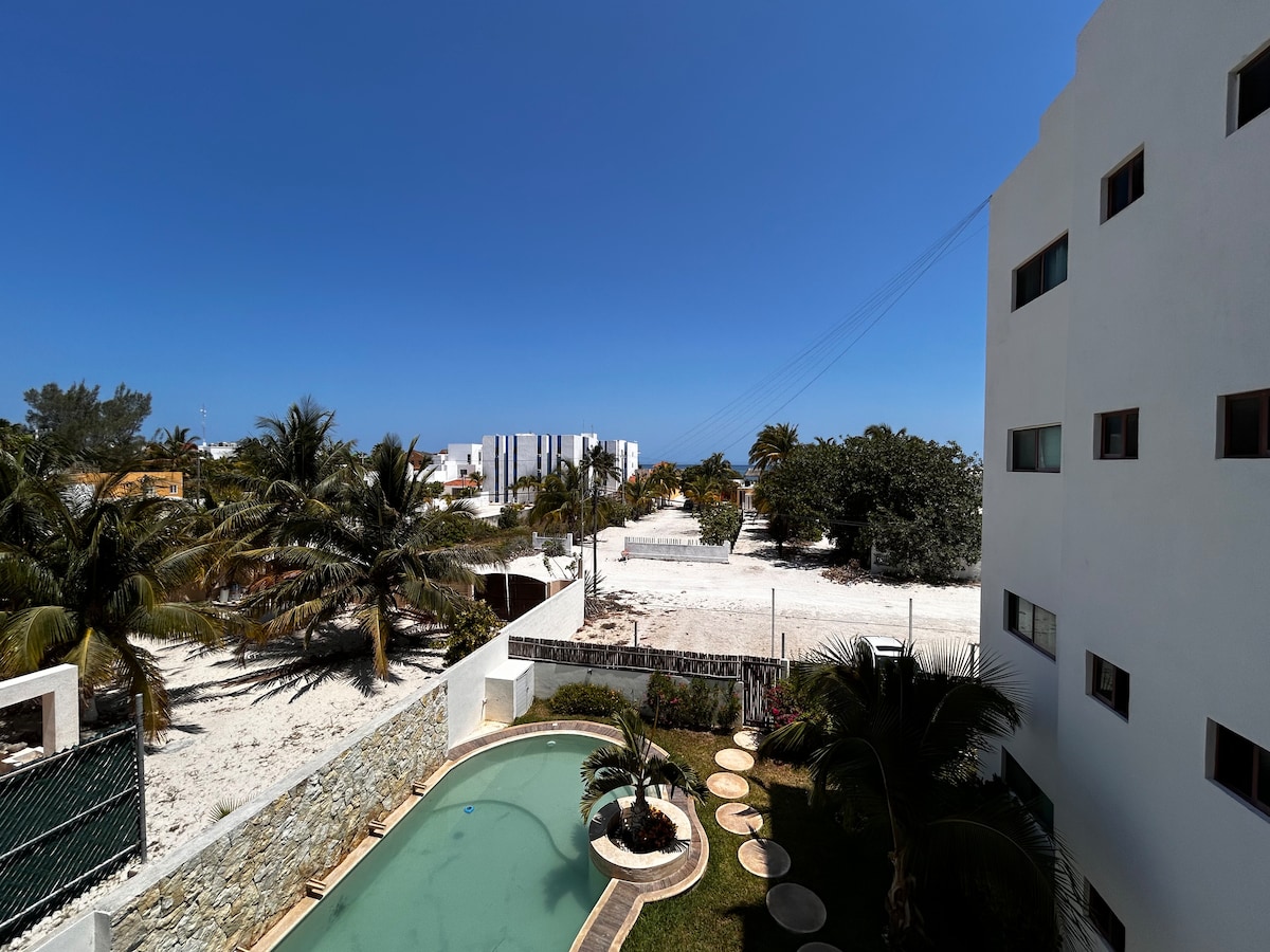 A clear view of the outdoor area is presented, featuring a swimming pool surrounded by lounge chairs. Palm trees line the property, and a sandy expanse is visible in the background. The bright blue sky enhances the inviting atmosphere of the space.
