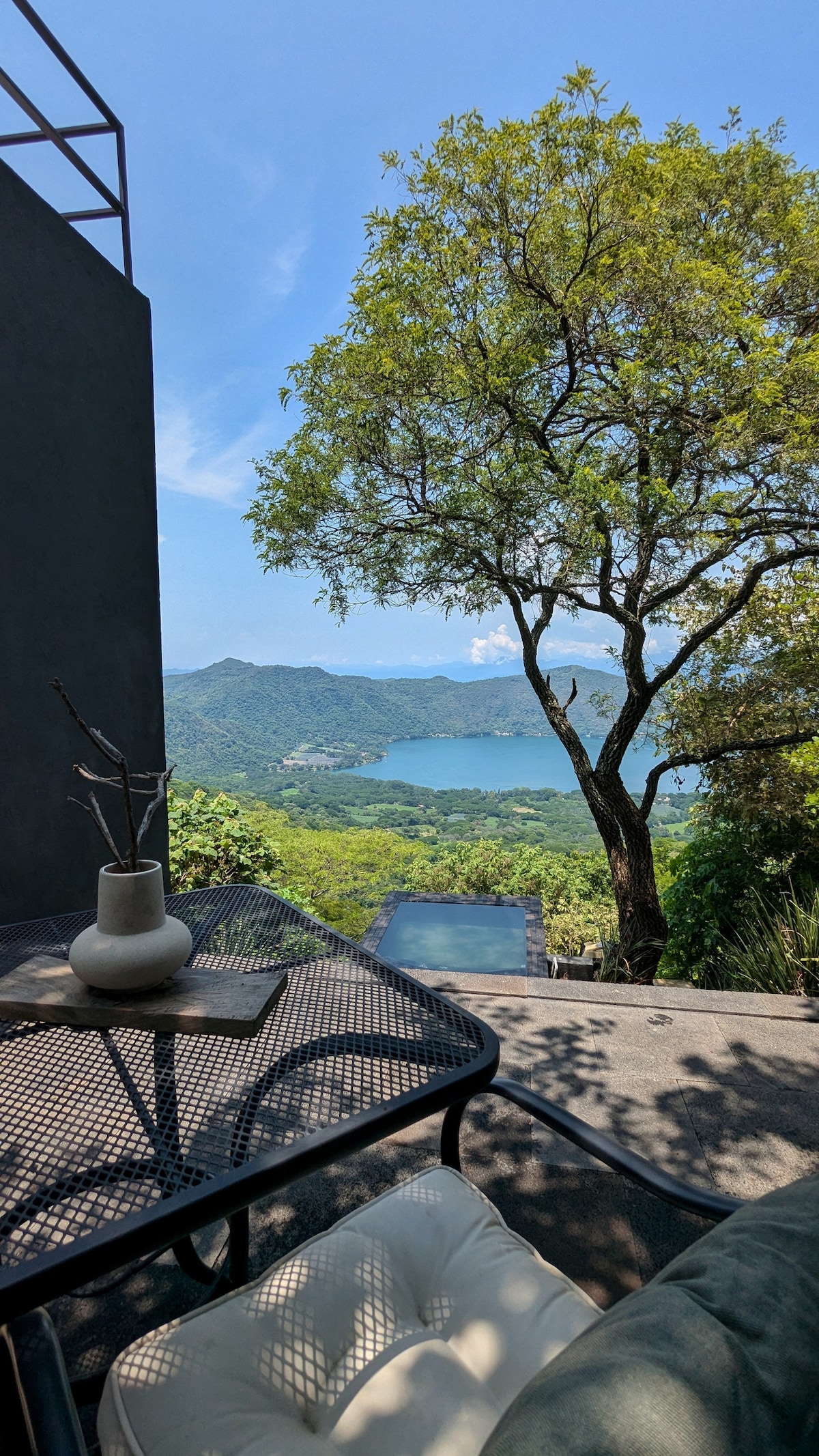 A terrace view showcases the sprawling landscape of Santa María del Oro lagoon, framed by a large tree and mountains in the distance. A glass table with a small vase is positioned on the patio, inviting relaxation while enjoying the natural surroundings.