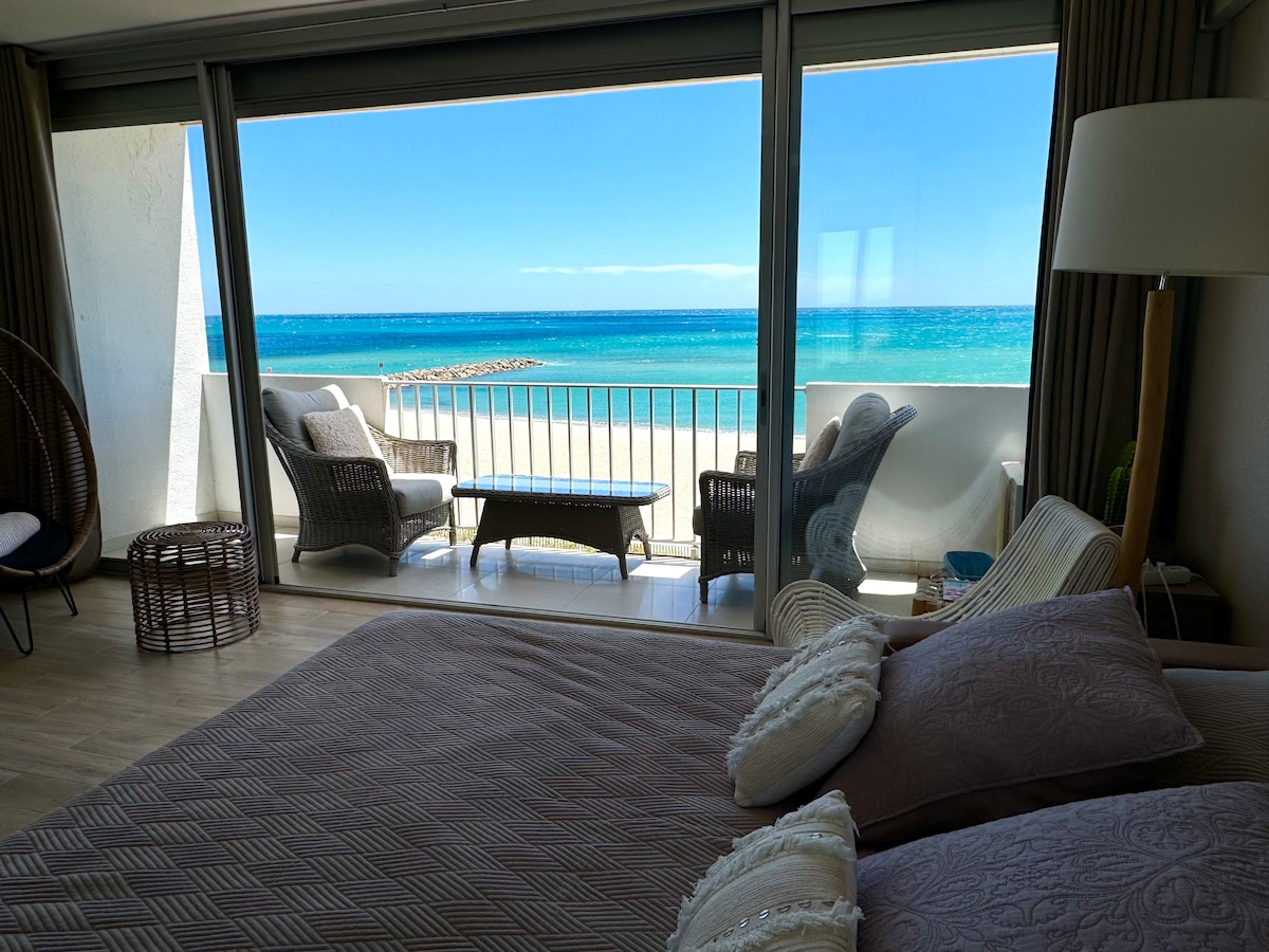 A spacious bedroom is presented, featuring a large bed with plush bedding. The panoramic sliding doors open to a private balcony overlooking the beach and ocean. Outdoor seating with comfortable chairs and a table is visible, inviting relaxation with stunning water views.
