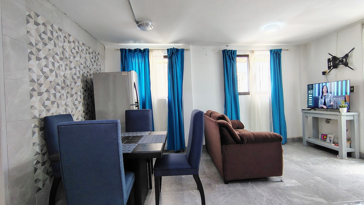 A modern living space features a dark brown sofa and a dining table with blue upholstered chairs. Large windows with blue curtains allow natural light to fill the room. A smart TV is positioned on a white console, accompanied by a refrigerator visible in the background.