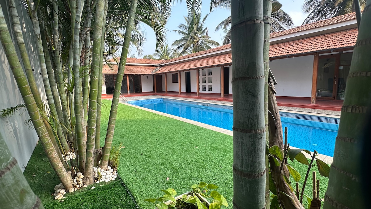 A private swimming pool is set against a backdrop of palm trees and lush greenery. The villa's spacious layout is visible, with rooms arranged along the perimeter. The pool reflects the sky, highlighting the tranquil atmosphere of the outdoor space.