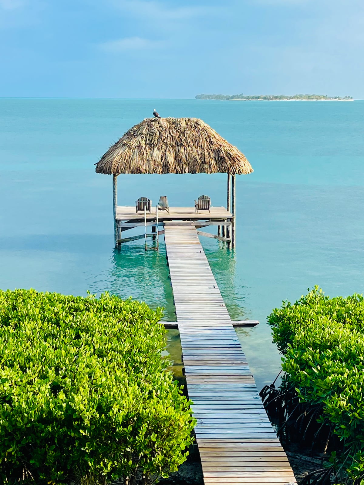 A wooden dock extends over calm turquoise waters, leading to a thatched-roof gazebo. Two chairs are positioned on the platform, surrounded by lush green mangroves, with a distant view of the coastline under a partially cloudy sky.