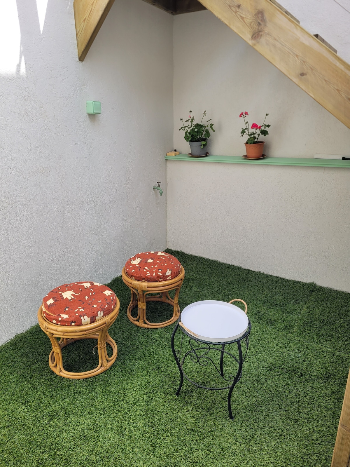 A small outdoor space is depicted, featuring two rattan stools with vibrant cushions. A round table stands between them. Green artificial grass covers the ground, with potted plants visible on a shelf above.