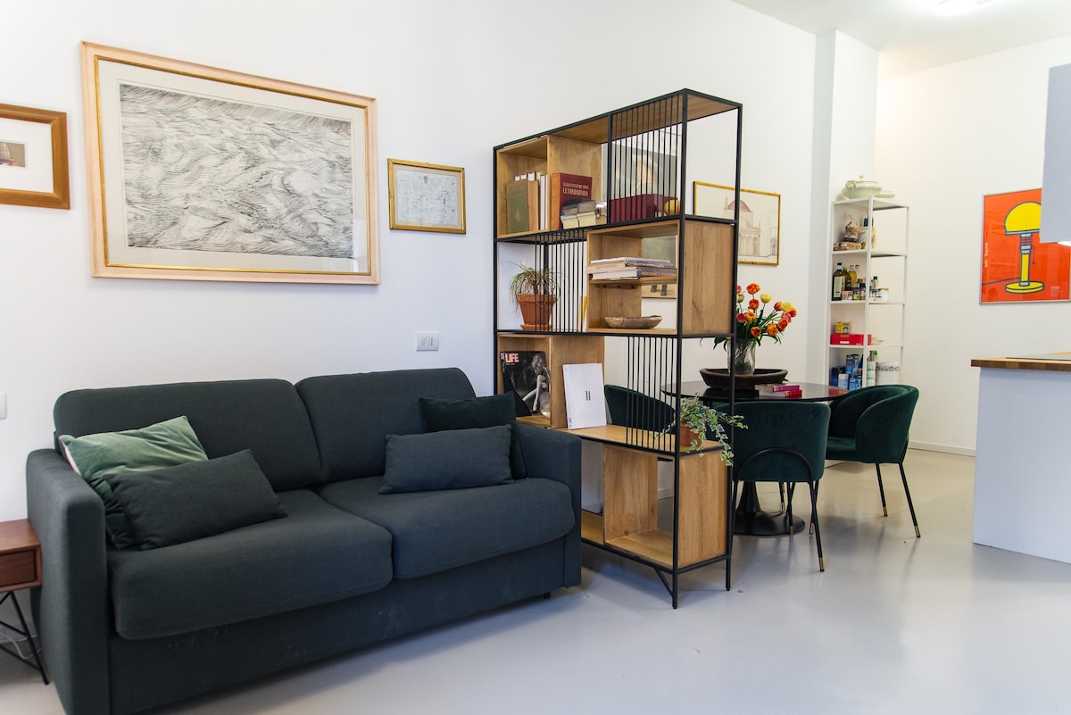A spacious living area features a comfortable dark green sofa with decorative pillows. A stylish bookshelf made of wood and metal stands nearby, filled with books and decorative items. In the background, a dining area with green chairs and a vibrant artwork adds a touch of modern design.