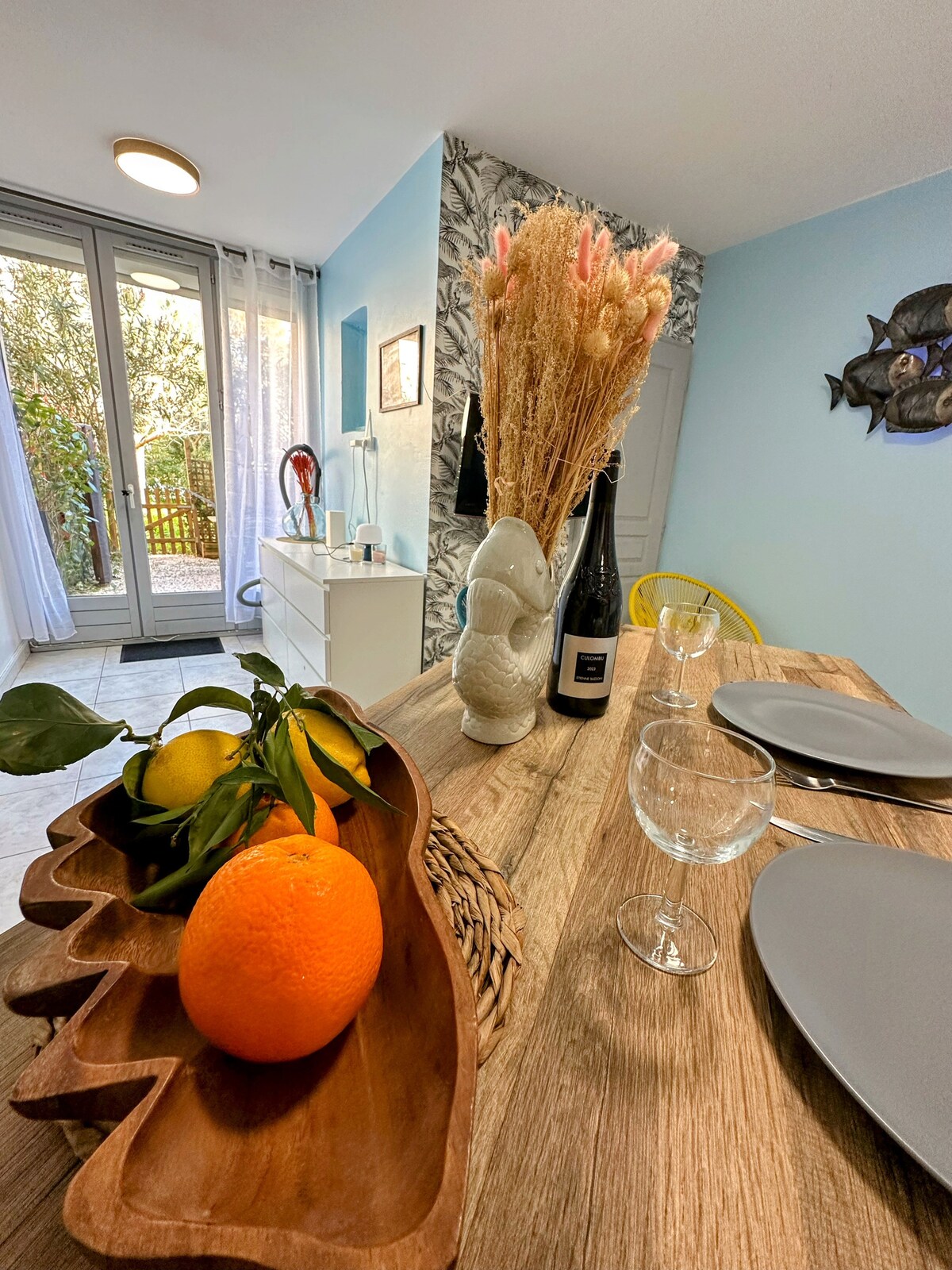 A dining area is presented with a wooden table set for two, featuring gray plates and glasses. A decorative bowl holds oranges and lemons. In the background, large glass doors open to a small outdoor space, with soft light filtering through.
