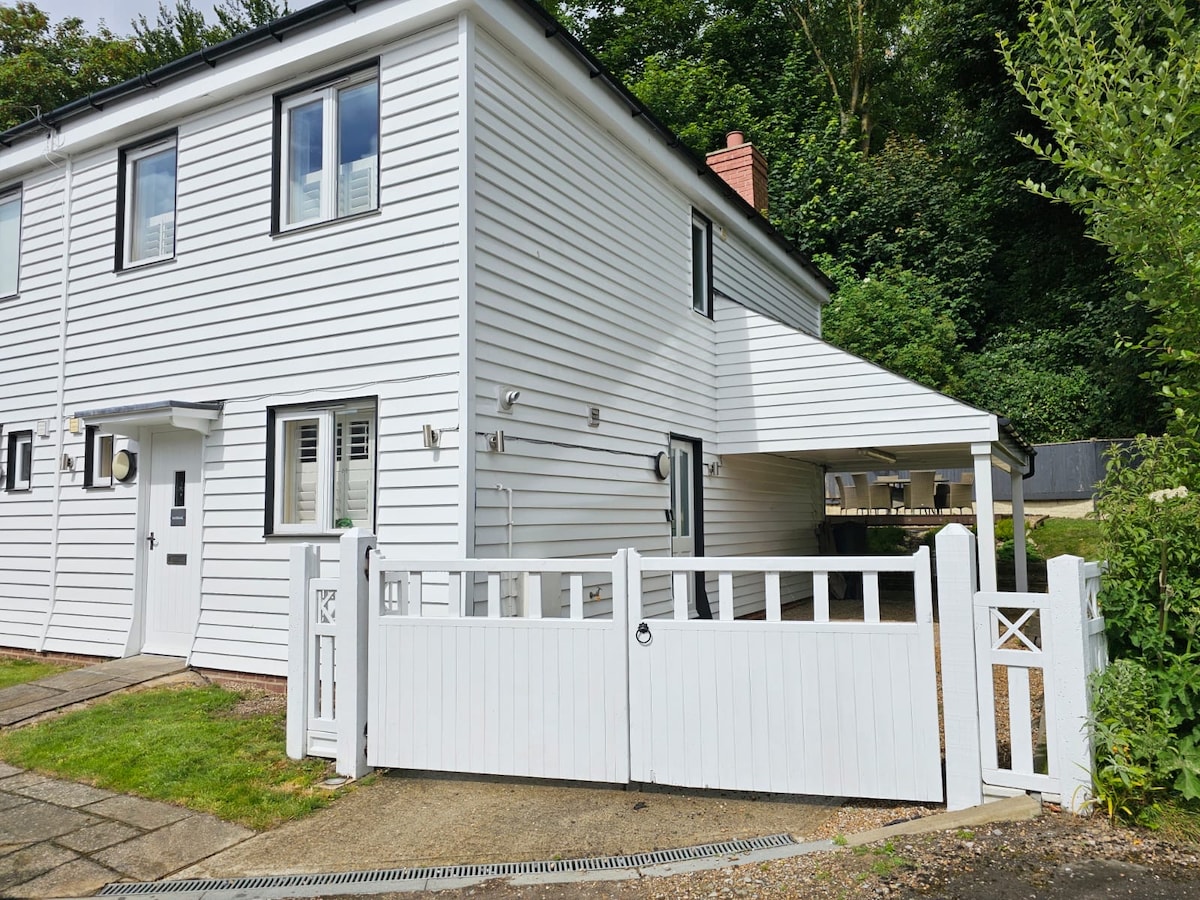 A modern cottage with a white wooden exterior is featured. The image shows a private gated entrance and a spacious driveway. Lush greenery can be seen in the background. A covered area extends from the side of the building, providing additional outdoor space.