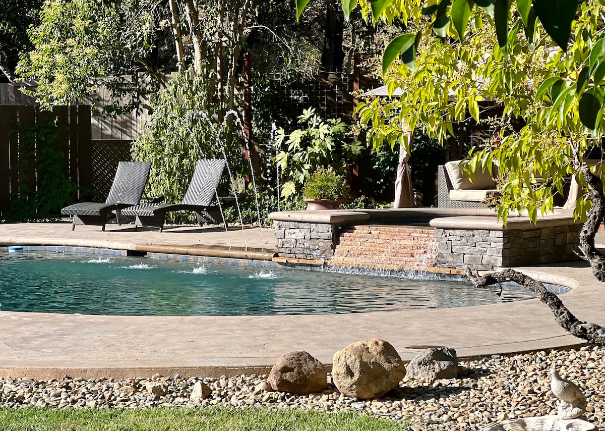 A serene outdoor area features a saltwater pool surrounded by a spacious stone deck. Two lounge chairs are positioned nearby. A cozy seating area is integrated into the stonework, with greenery and natural stones adding to the tranquil setting.