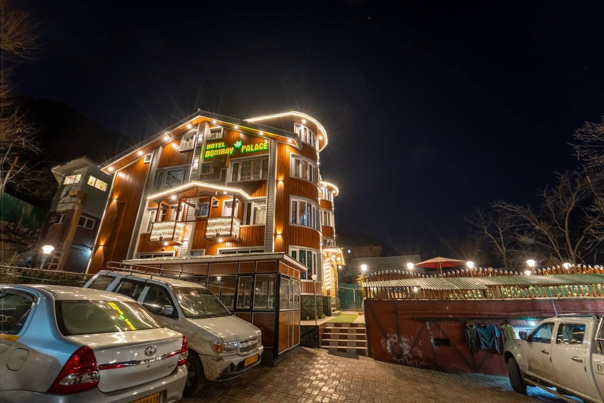 The exterior of Bombay Palace is showcased at night, featuring warm lighting that highlights its wooden architectural details. The entrance is framed by lush greenery, while several parked cars are visible in front of the building. The inviting ambiance enhances the welcoming appeal of the hotel.