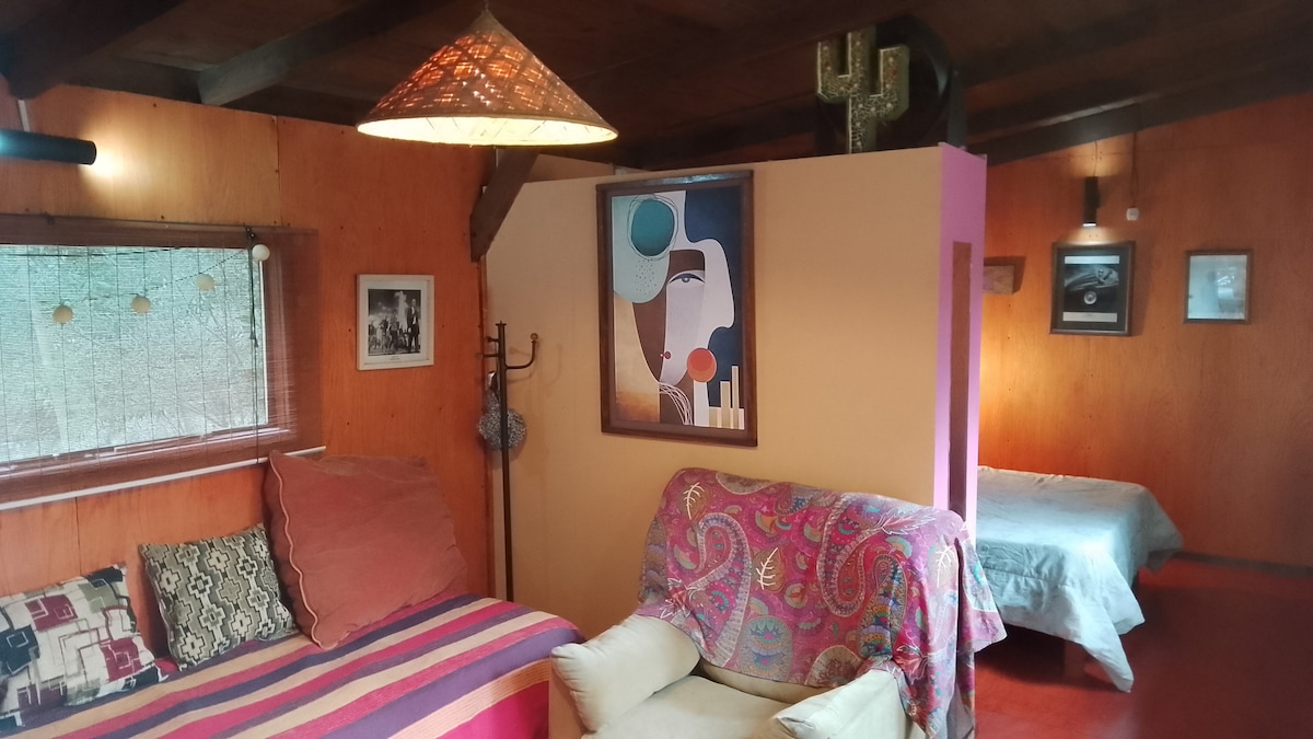 An inviting living area features a colorful tapestry draped over a comfortable chair, with a striped orange and pink sofa beside it. A prominent artwork is displayed on the wall, and soft lighting is provided by a hanging lamp. A spacious bed is positioned toward the back.