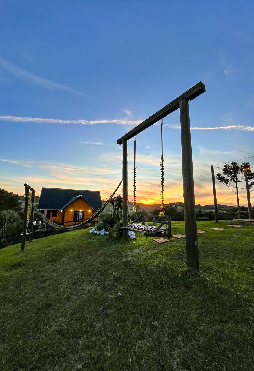 A wooden swing set is positioned on a grassy area, overlooking a vibrant sunset. The backdrop features a cozy chalet with a dark roof, partially illuminated by the warm glow of the setting sun. Trees add to the peaceful landscape.
