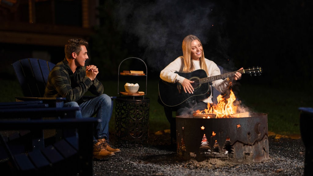 Around a fire pit, two individuals are seated on Adirondack chairs. One person plays an acoustic guitar while the other listens attentively. The firelight casts a warm glow, creating a cozy atmosphere in the outdoor setting.