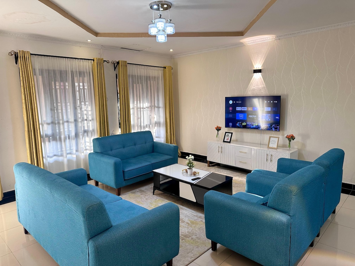A spacious living area is furnished with two blue sofas facing each other, complemented by a modern coffee table. A large television is mounted on the wall, and curtains frame the windows, allowing natural light to fill the space. Decorative items, including a small vase, add a touch of warmth.