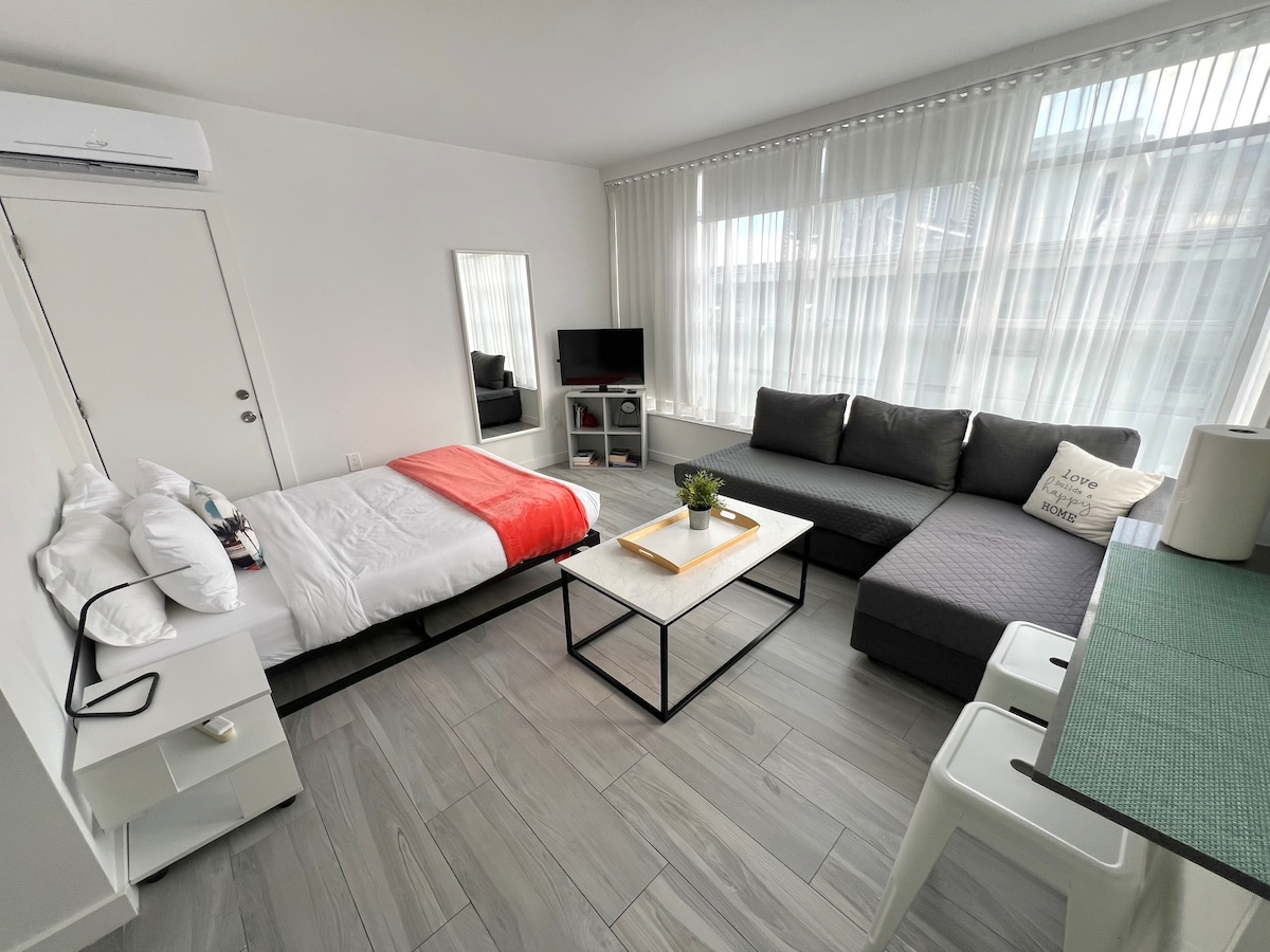 A well-appointed studio apartment is presented, featuring a spacious bed with crisp white linens and a vibrant orange throw. A cozy sectional sofa complements a sleek coffee table. Natural light filters through sheer curtains, creating a bright and airy ambiance. A mirror and TV add functionality to the space.