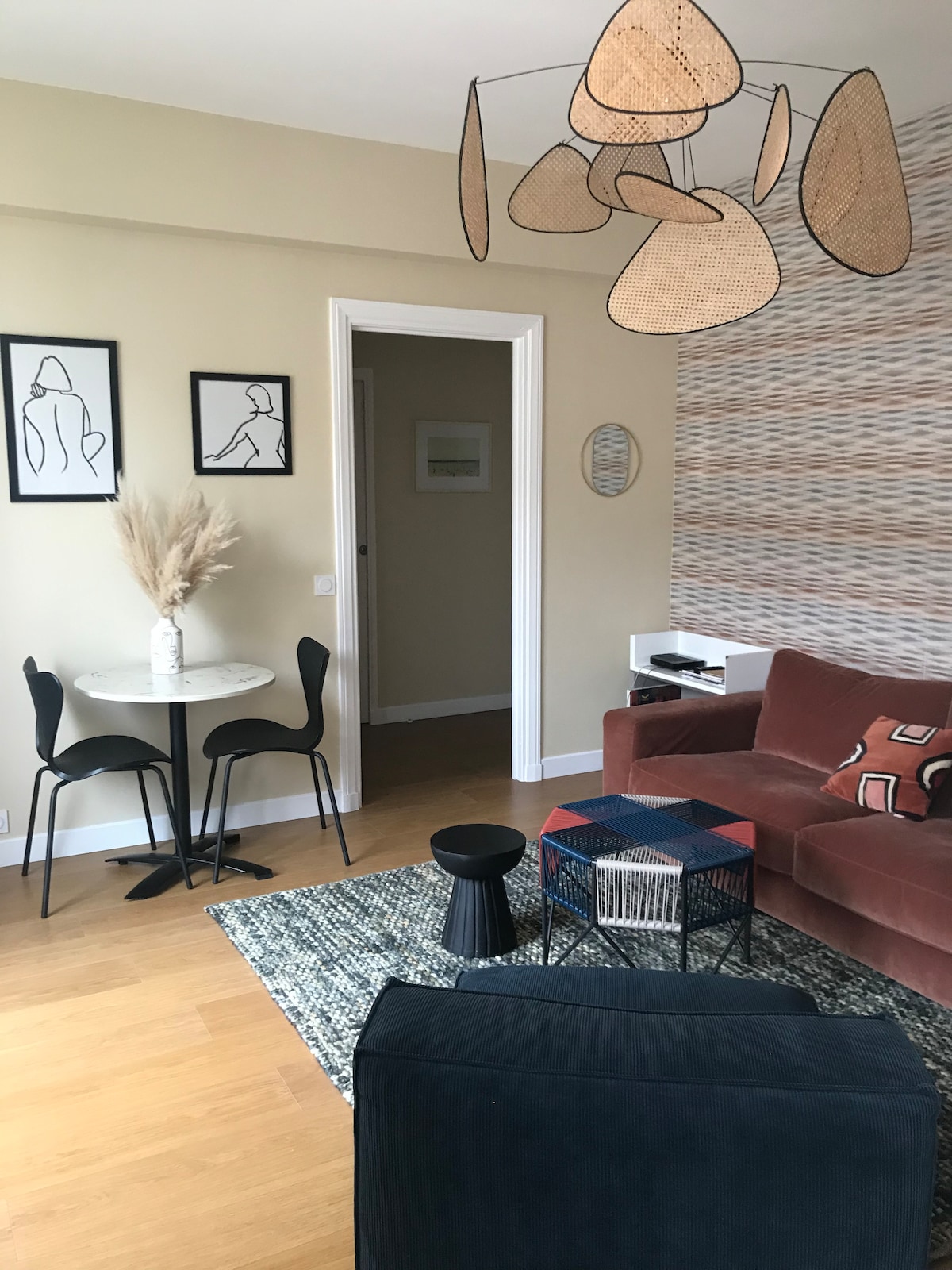 A bright living area features a comfortable maroon sofa adorned with decorative cushions. A round dining table with two chairs sits nearby. The space is enhanced by abstract wall art and a unique hanging light fixture, creating an inviting atmosphere with natural light coming through a doorway.